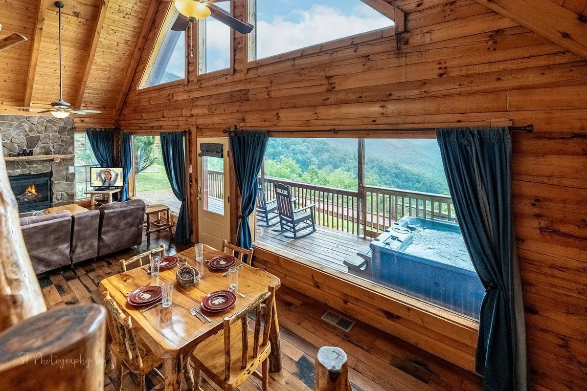 A spacious, light-filled area with large windows looking out over scenic vistas, a rustic dining table, a comfortable seating area by a fireplace, and a hot tub visible on the deck. The ambiance speaks to shared moments, warmth, and a seamless connection to the surrounding landscape.