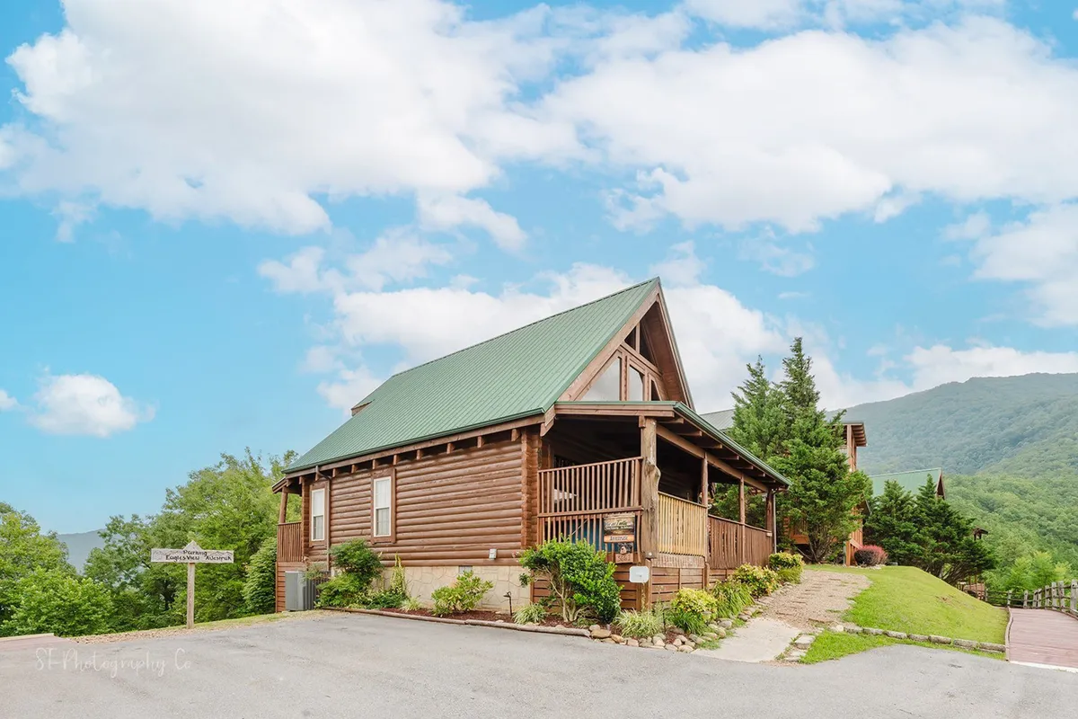 A classic log cabin with a green metal roof is nestled among tall trees, with a dramatic mountain backdrop. The inviting exterior sets the tone for a secluded yet accessible adventure basecamp, surrounded by nature and framed by the forest and sky.