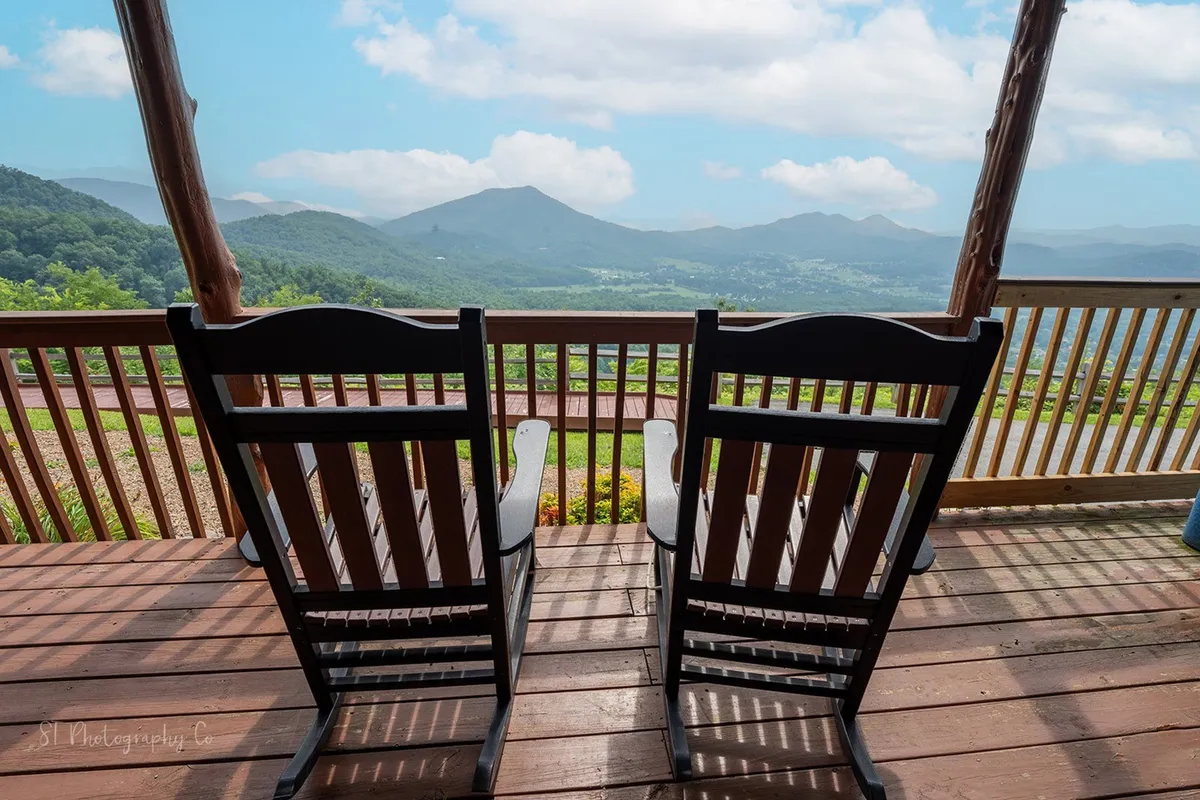 A tranquil scene of the deck with a pair of classic wooden rocking chairs facing outward over a mountain vista, framed by the deck railing and surrounded by lush greenery. The peaceful atmosphere is ideal for reading, conversation, and soaking in the sunrise or sunset.