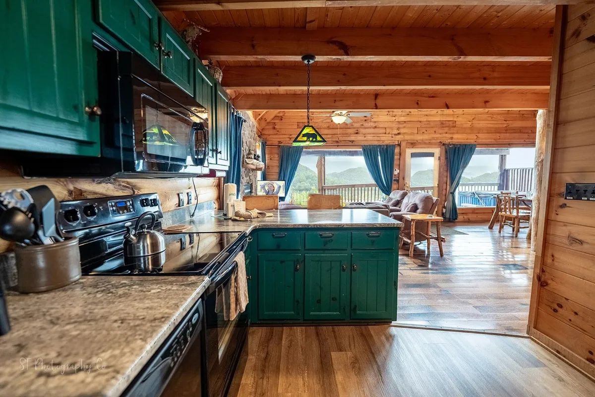 Featuring green kitchen cabinets, stainless steel appliances, wooden beams, and a large window with a scenic view, this image captures the inviting and modern aspect of the kitchen. The breakfast bar and ample seating are subtly visible, reinforcing the idea of a communal and versatile space.
