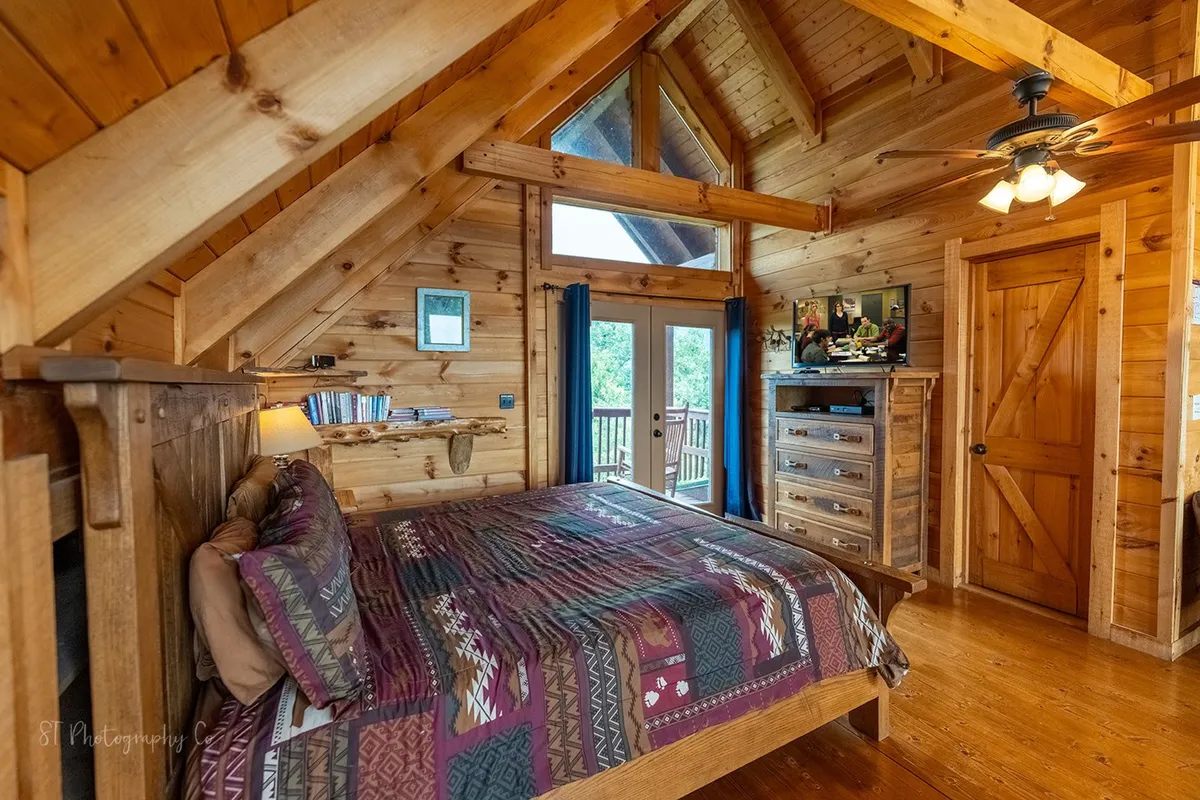 A wooden bed with colorful bedding sits beneath large windows that flood the room with natural light. The room is equipped with a television unit and wooden furniture, creating a warm, inviting space perfect for relaxation and privacy.