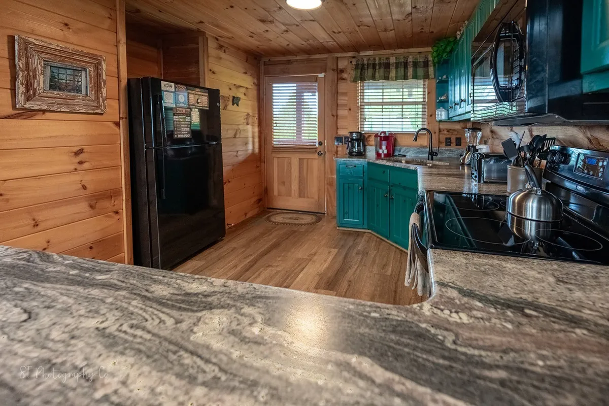 This image features a warm, inviting kitchen with teal cabinets, granite countertops, and modern appliances, set against wooden walls. The scene suggests a space perfect for family gatherings, preparing home-cooked meals, and creating lasting memories together.