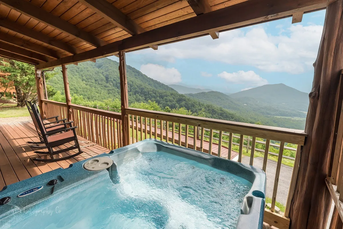 An inviting exterior shot of the deck featuring a bubbling hot tub, two wooden rocking chairs, and sweeping mountain views beyond the railing. The scene captures a tranquil moment at sunrise, with steam rising from the hot tub and soft natural light illuminating the porch.