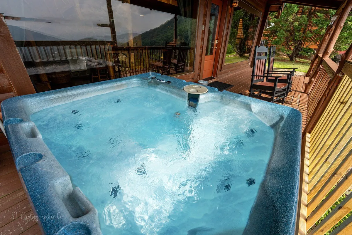 A bubbling hot tub sits on a spacious wooden deck, surrounded by lush greenery and offering sweeping views of the hills and distant mountains. The tranquil scene conveys relaxation, with room for friends or family to gather and enjoy the sunset or a soothing soak after a day outside.