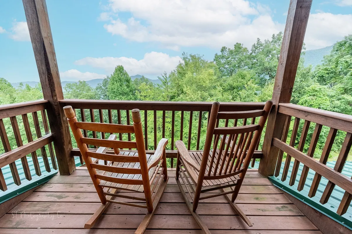 This exterior image features a pair of wooden rocking chairs on a deck, overlooking lush greenery and expansive mountain views. The setup suggests a serene space for couples or friends to relax, converse, and enjoy the scenic beauty day or night.