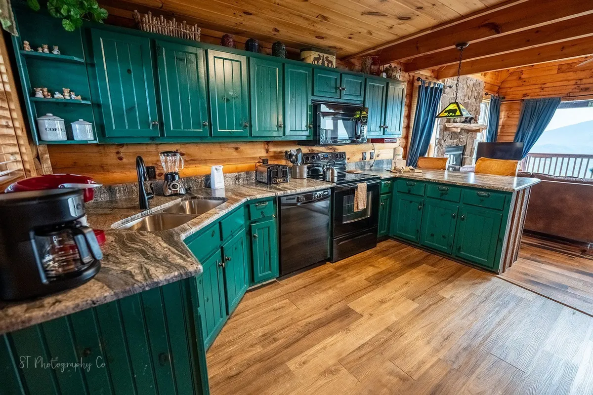 This image showcases the kitchen with vibrant green cabinetry, granite countertops, wooden beams, and large windows that offer ample natural light. The space is welcoming and designed for gatherings, with enough room for meal preparation and family interaction.