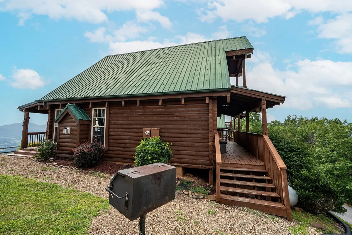 The image shows a classic log cabin with a green metal roof, a wooden deck, and an outdoor grill, all set against a backdrop of lush greenery. The deck invites gatherings, barbecues, or simply relaxing with a view of the Smoky Mountains.