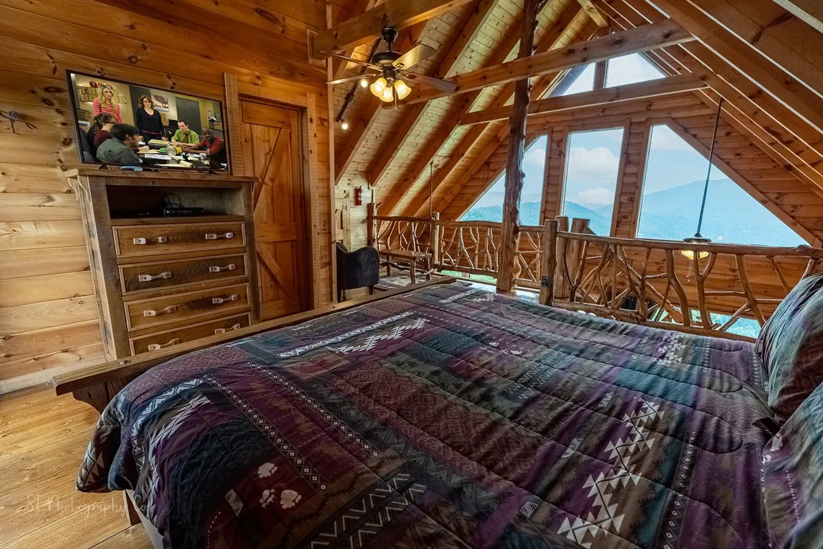 This image showcases a cozy bed with a patterned quilt positioned next to large windows that offer sweeping mountain views. The wooden furniture and tranquil atmosphere are enhanced by a TV, inviting guests to relax and unwind after a day outdoors.