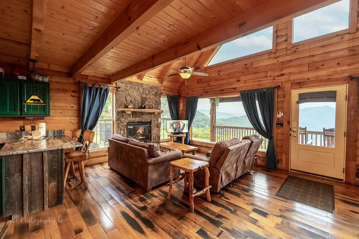 An open-concept living area featuring a striking stone fireplace, ample rustic wooden furniture, and large windows with breathtaking mountain vistas. The kitchen area with green cabinets is visible, illustrating the seamless flow between spaces.