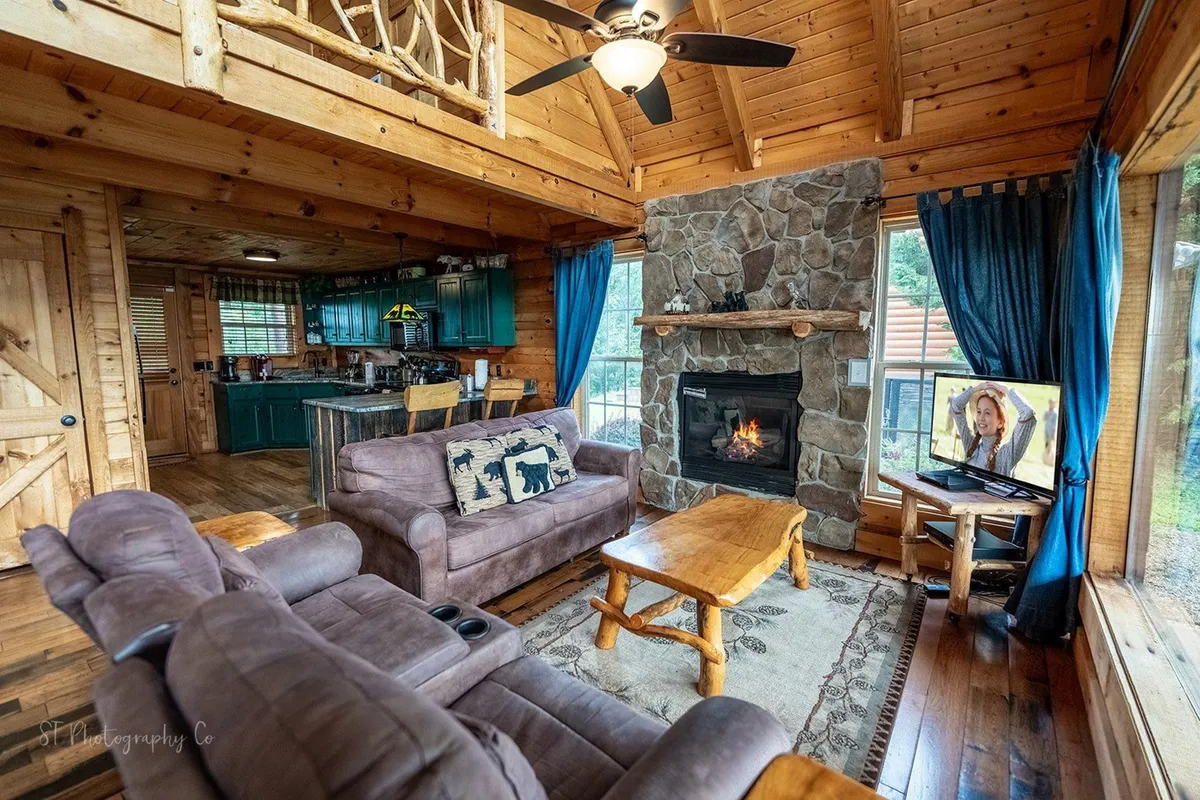 A comfortable living room scene with a stone fireplace, cozy couches, rustic wooden furniture, and large windows. The television and a generous seating arrangement invite gatherings and movie nights.