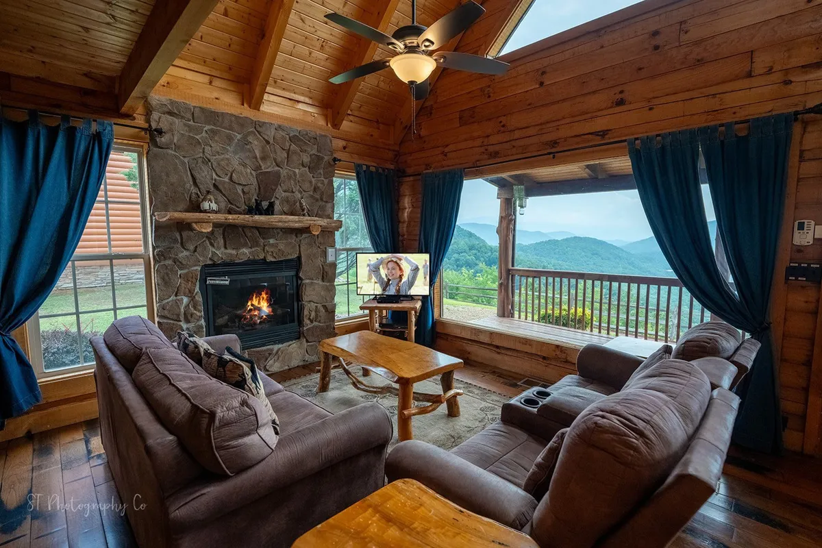 A welcoming living room with a stone fireplace as the focal point, flanked by large windows offering mountain views. Plush sofas and a coffee table are arranged for relaxation, with a flat-screen TV mounted nearby. The space is flooded with natural light, and the open layout hints at seamless flow to adjoining areas.