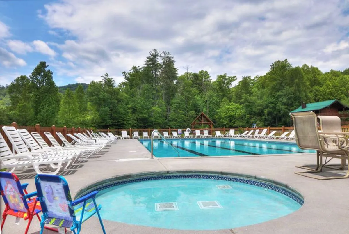 A cheerful outdoor pool scene featuring a sparkling swimming pool, adjacent hot tub, comfortable lounge chairs, and lush surrounding trees. The space is designed for both kids and adults to swim, sunbathe, and relax in a family-friendly environment.